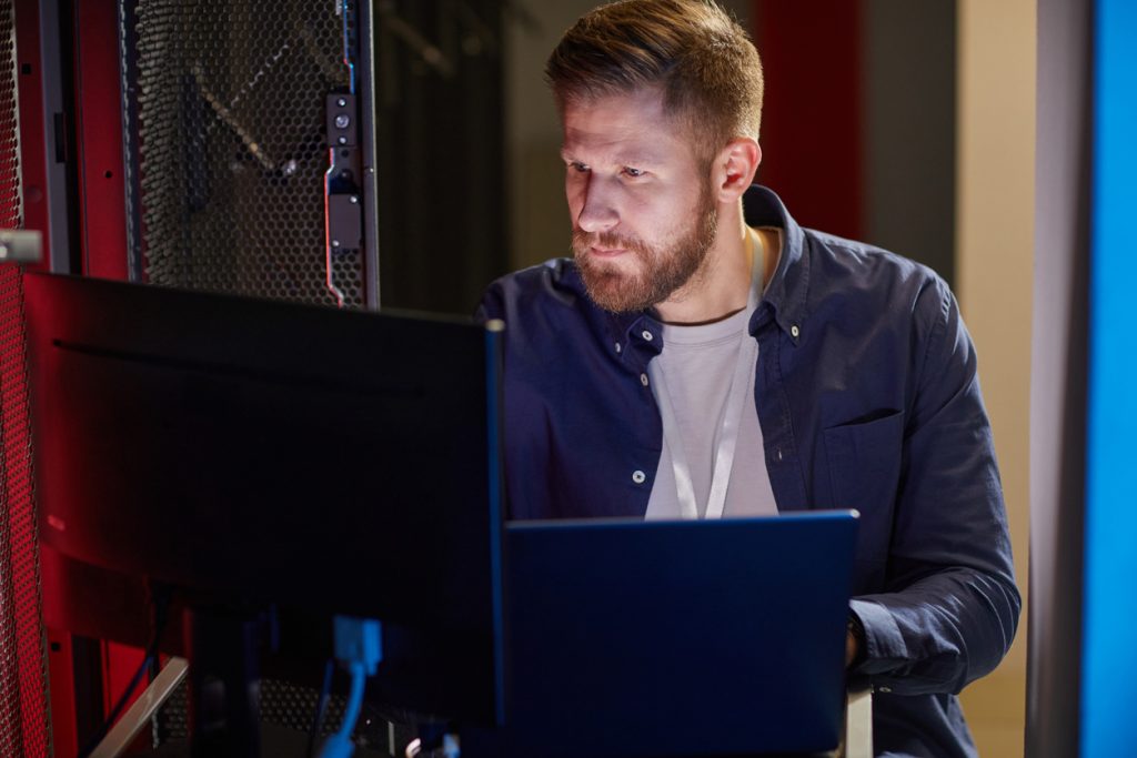 Male IT Professional looking at two monitors attached to a server rack.